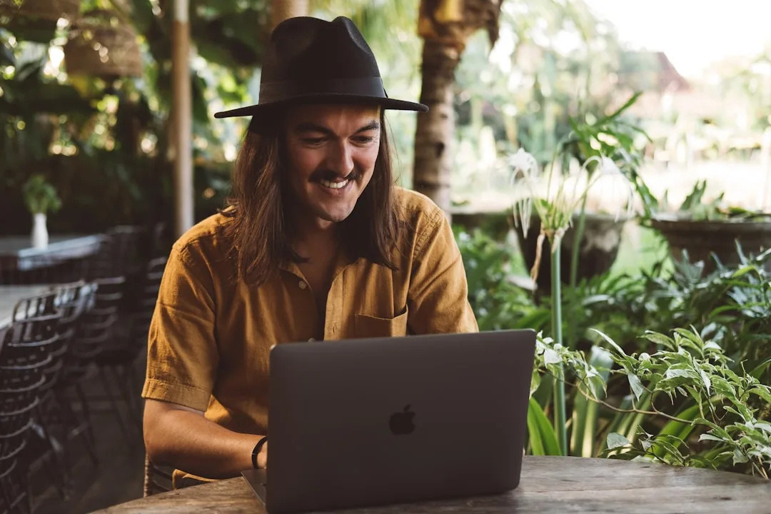 Mann mit schwarzem Hut arbeitet lächelnd am Laptop in einem tropischen Gartenlokal