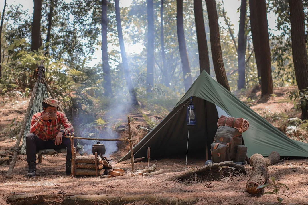 Camper sitting outside a green tent in sunlit forest with camping gear around
