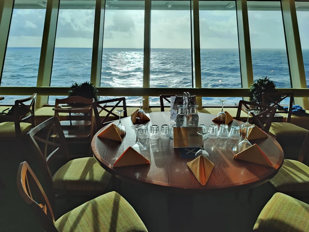 Formell gedeckter Dining-Tisch mit Weingläsern an der Panorama-Glasfront eines Kreuzfahrtschiffs mit Meerblick